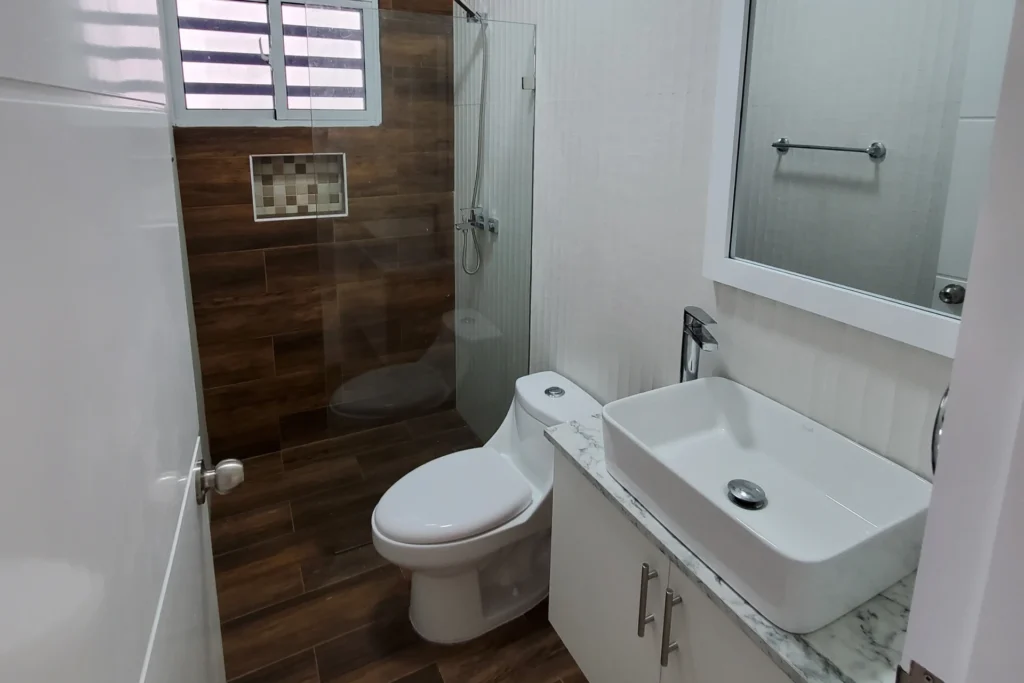 Baño con lavabo rectangular, inodoro y ducha con mampara transparente y pared de tonos marrones.