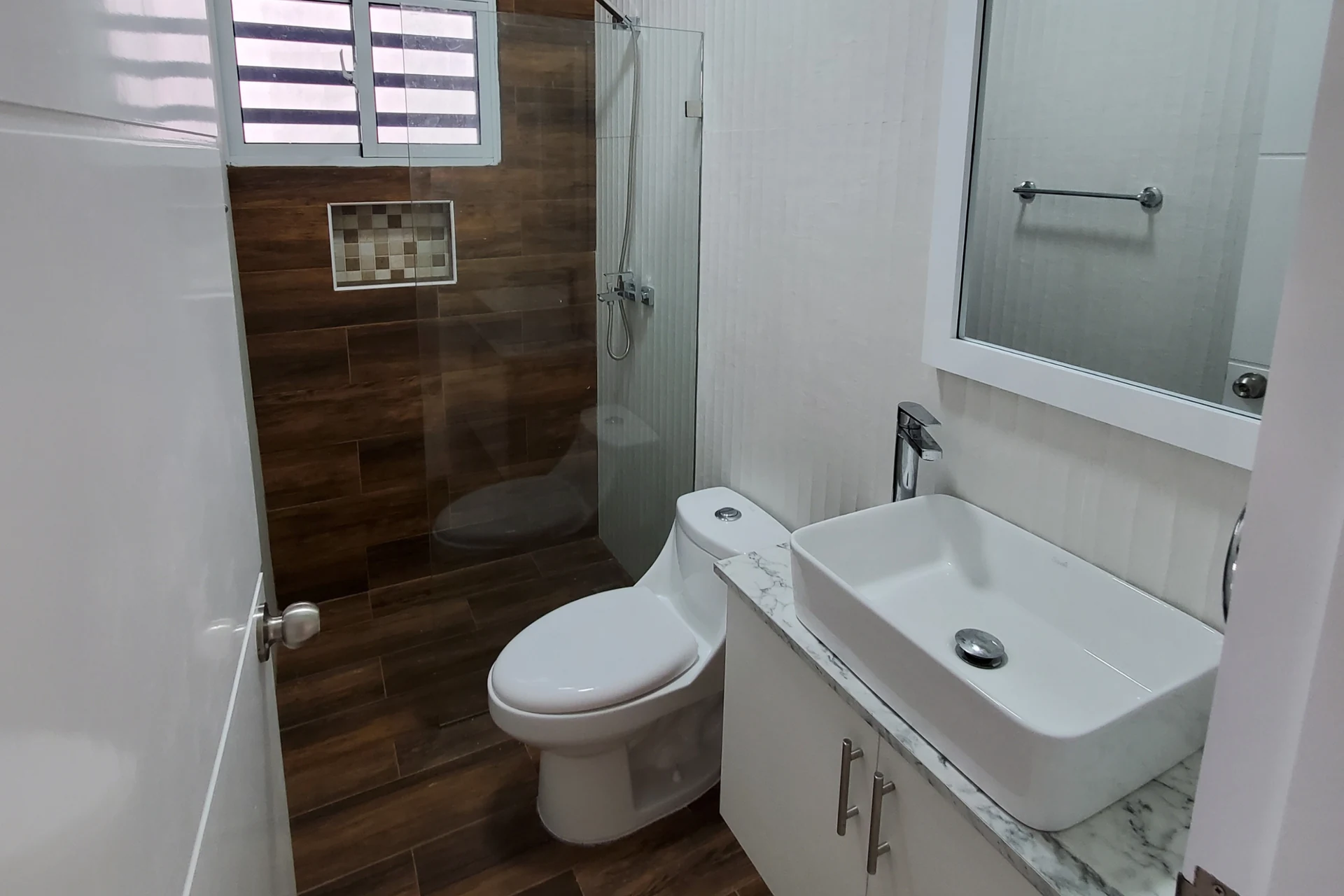 Baño con lavabo rectangular, inodoro y ducha con mampara transparente y pared de tonos marrones.