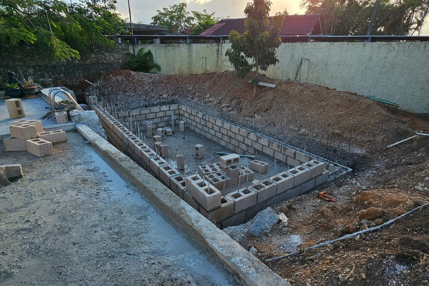 Excavación para una piscina con muros de bloques, materiales de obra y árboles junto al cerramiento.