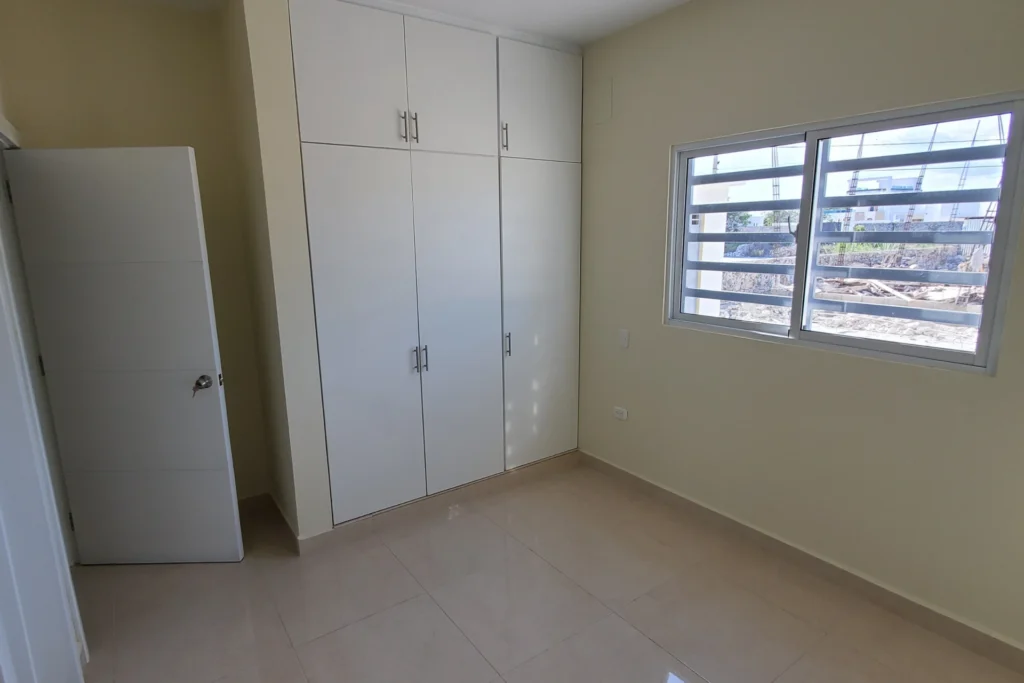 Habitación vacía con armario blanco de puertas altas y ventana con barras horizontales.