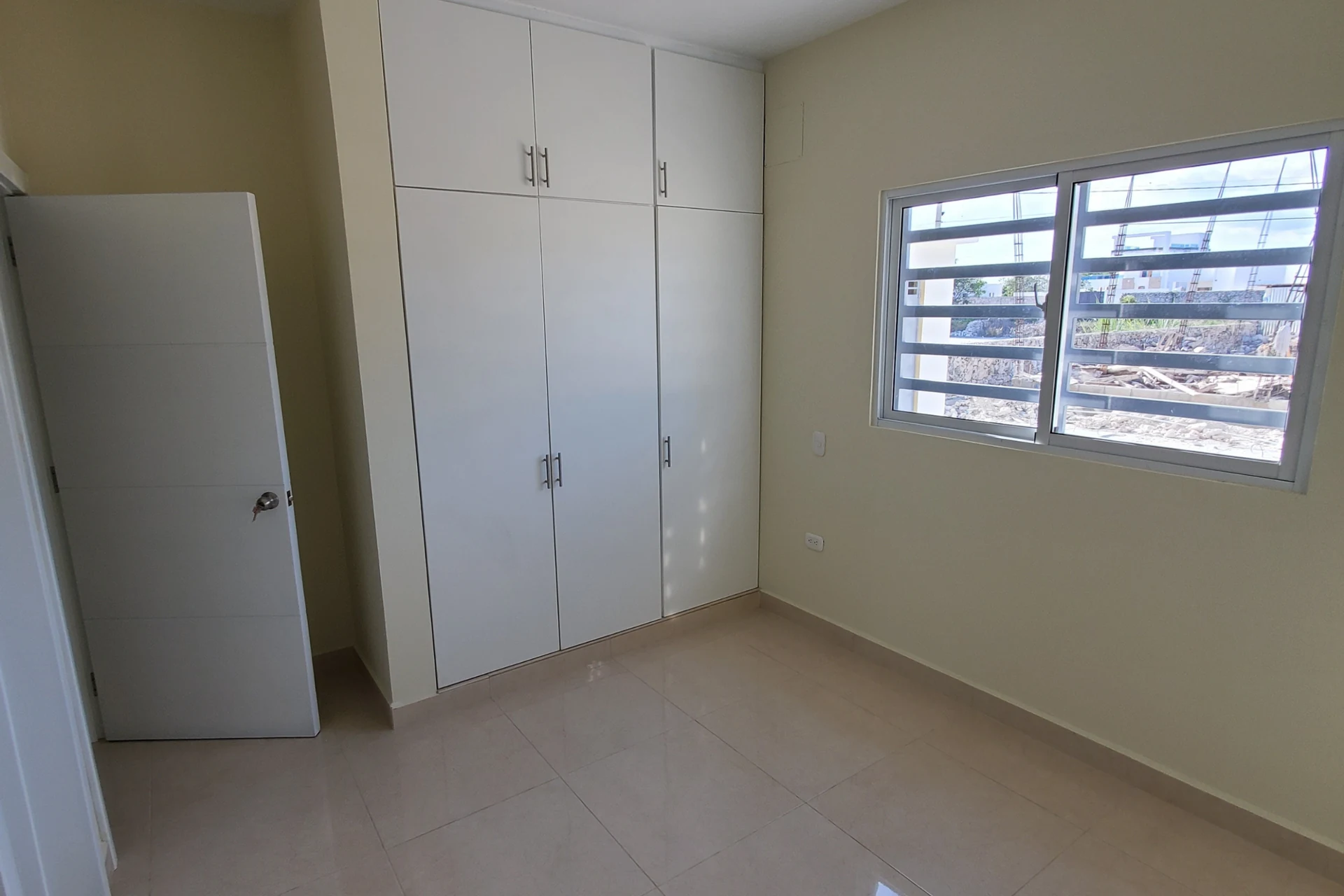 Habitación vacía con armario blanco de puertas altas y ventana con barras horizontales.