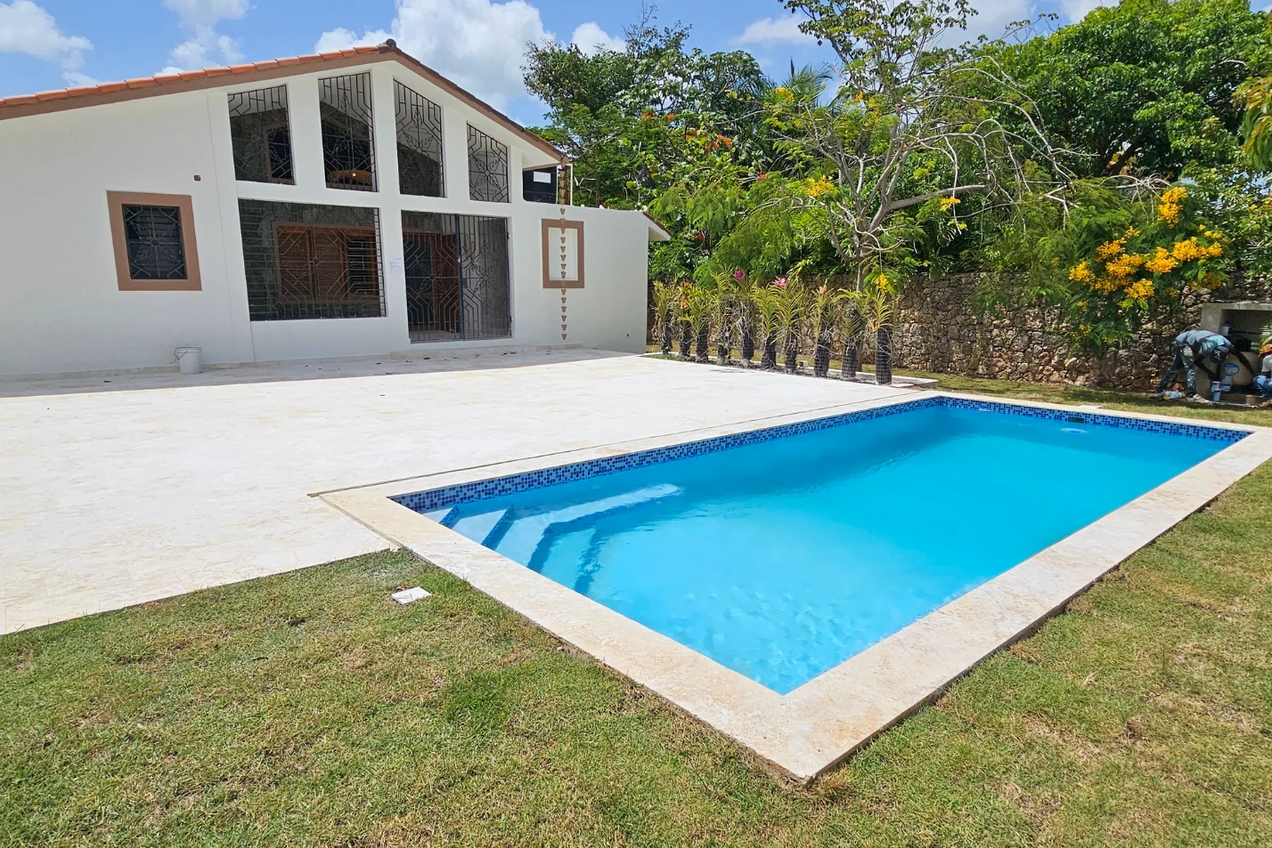 Piscina rectangular terminada junto a una vivienda blanca, una terraza pavimentada y césped.