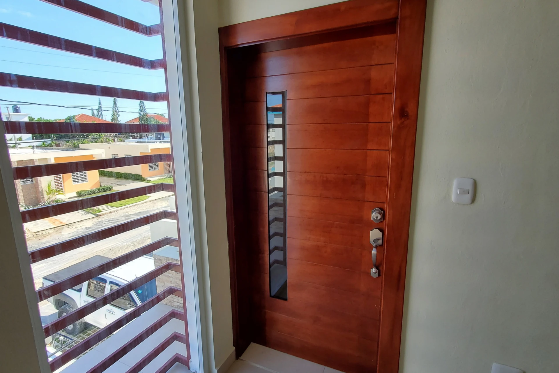 Puerta marrón con franja vertical de vidrio junto a un ventanal con listones horizontales rojizos.