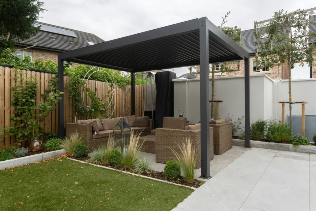 Terraza con pérgola negra, sillones de exterior y jardín junto a una vivienda.